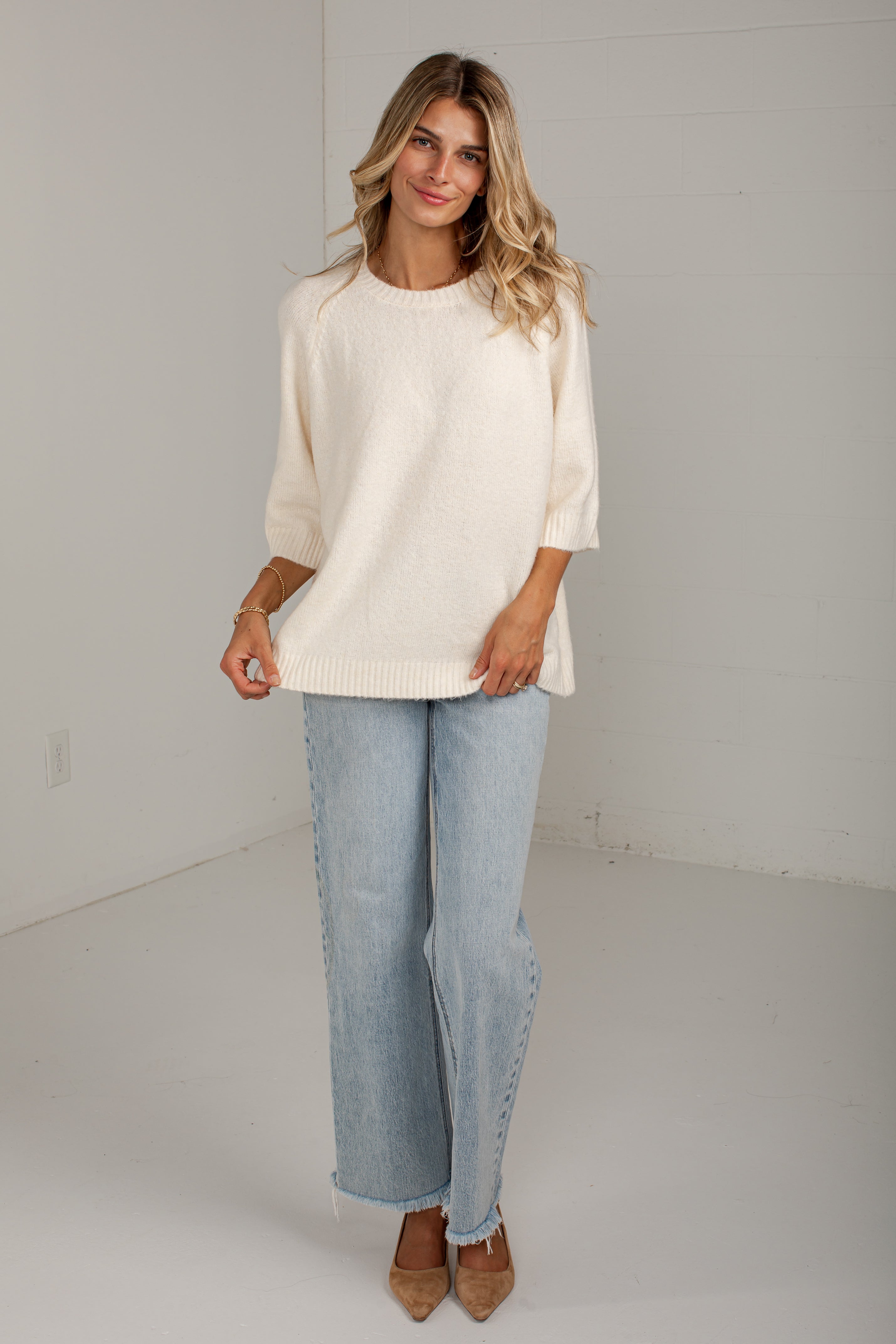 A woman modeling a cream-colored knit sweater with three-quarter sleeves, paired with light-wash wide-leg jeans and tan pointed-toe heels, standing against a white studio wall.