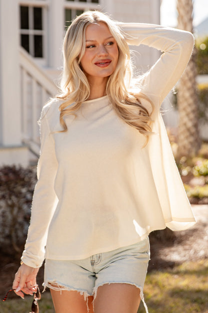 Woman in a white sweater and denim shorts standing outdoors with a house and palm tree in the background.