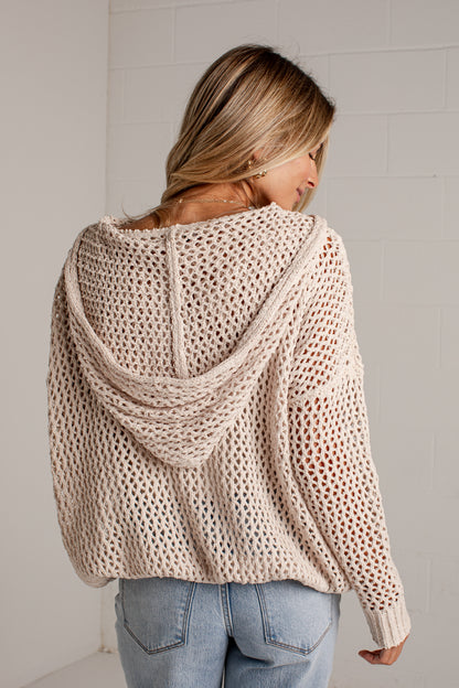 A woman viewed from behind wearing a chunky open-knit hooded sweater in a light beige color, paired with light-wash blue jeans, against a white brick wall.