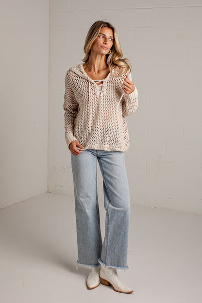 A woman modeling a cream-colored open-knit cropped sweater with a lace-up neckline, paired with light-wash wide-leg jeans and white ankle boots, standing in a minimalist studio setting.