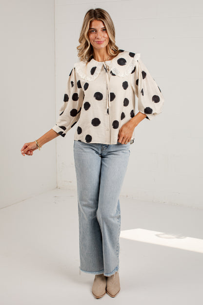 A woman modeling a cream-colored blouse with black polka dots, a large Peter Pan collar, and puff sleeves, paired with light-wash blue jeans and beige ankle boots, in a bright white studio setting.