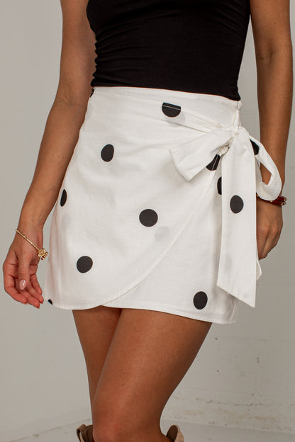 A woman wearing a white wrap-style mini skirt with black polka dots and a tied waist, paired with a black top against a plain light background.