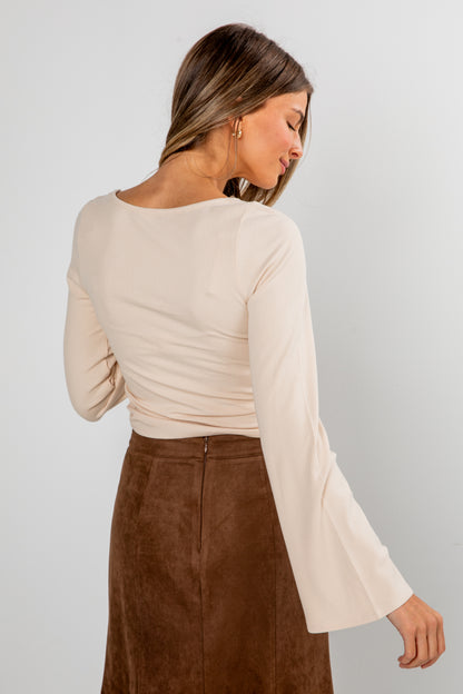 Woman wearing a beige long-sleeve top and brown skirt on a plain background