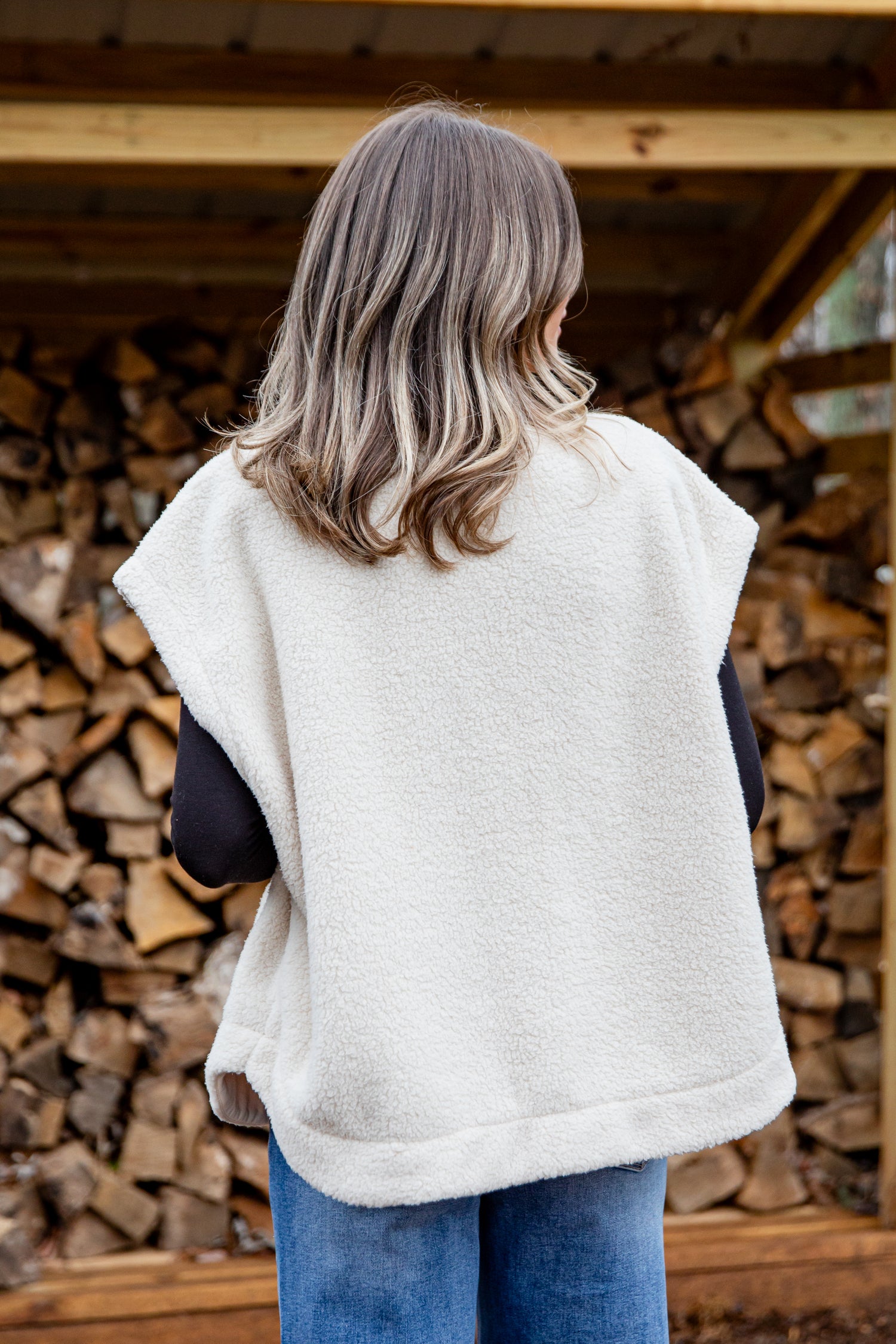 Person wearing a white vest standing in front of stacked firewood.