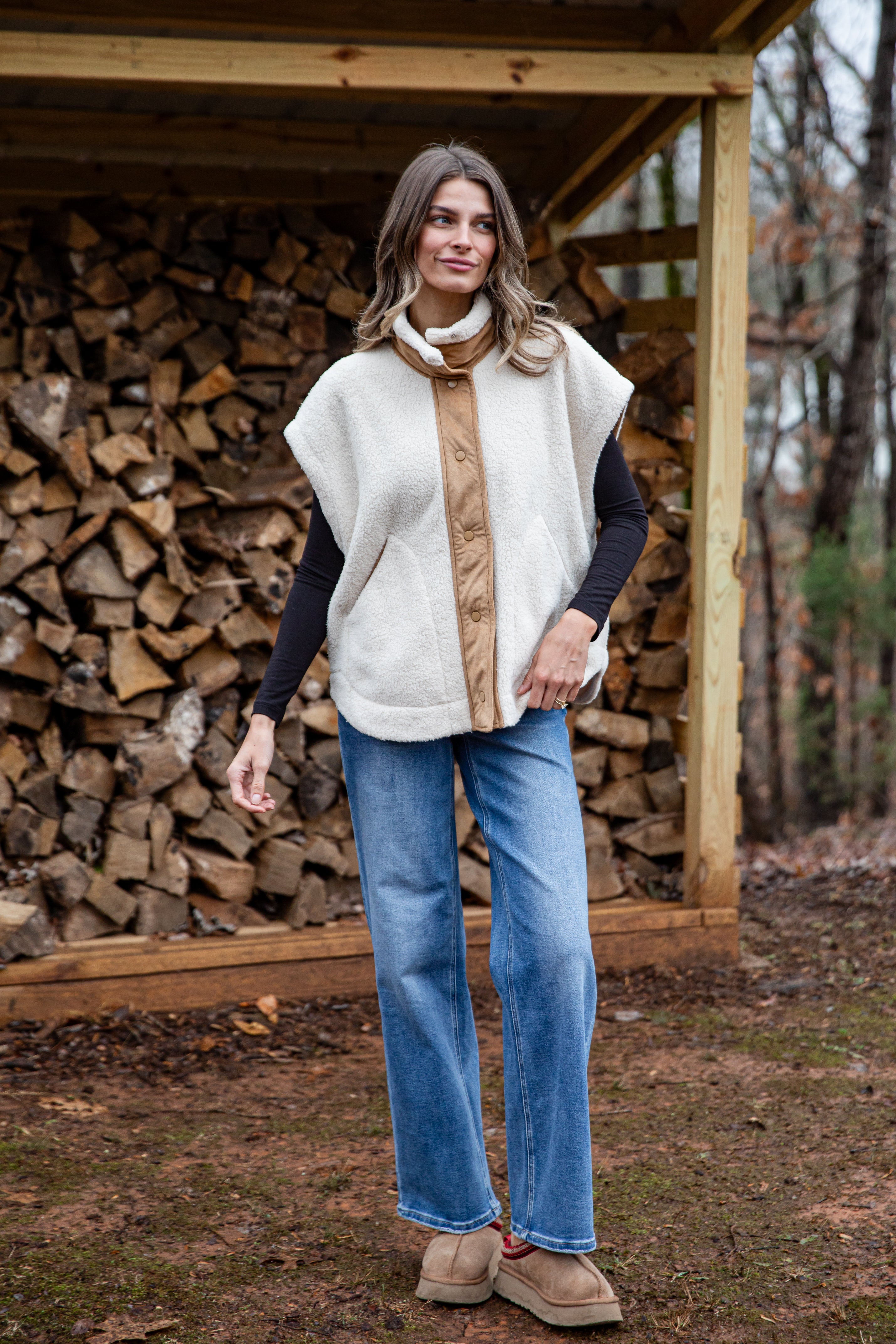 Woman wearing a white vest over a brown shirt with blue jeans, standing in front of a wooden structure with stacked firewood.