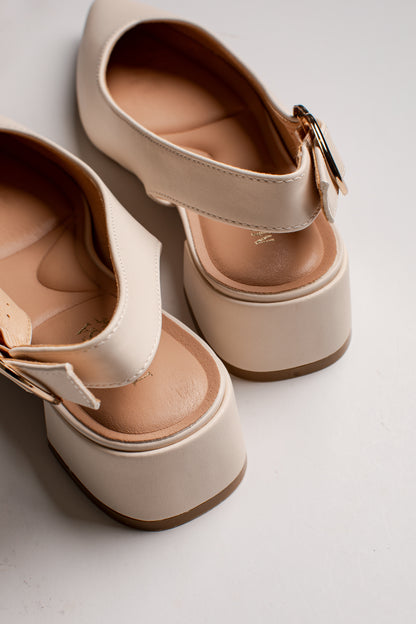 A pair of platform Mary Jane-style shoes in a warm cream or off-white color, featuring a single strap with a small gold-tone buckle and a tan insole, shown on a light gray background.
