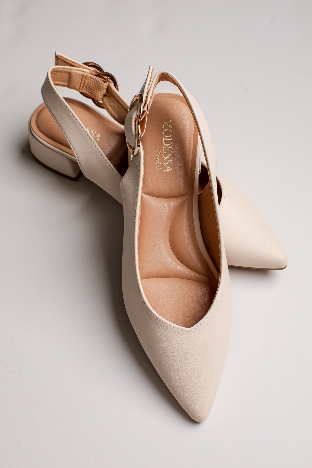 A pair of pointed-toe slingback heels in a light cream color with a gold-tone buckle, displayed on a light gray background. The brand name &