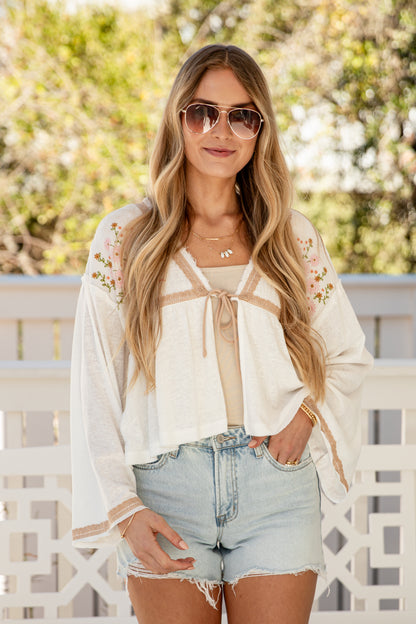 Woman wearing a white blouse with floral embroidery and denim shorts outdoors.