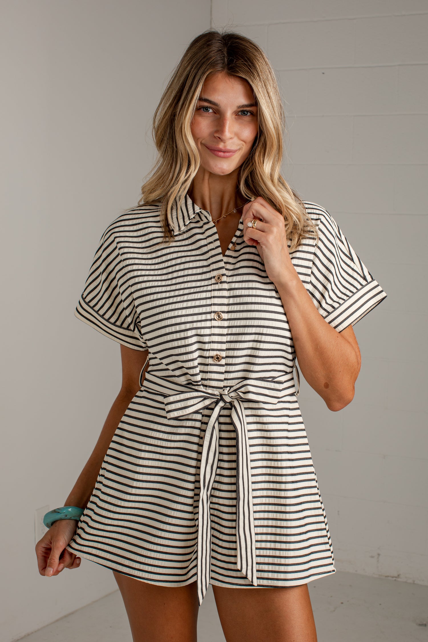 A woman modeling a short-sleeve collared romper with thin black horizontal stripes on a cream background, featuring a button-front bodice and a self-tie waist belt.