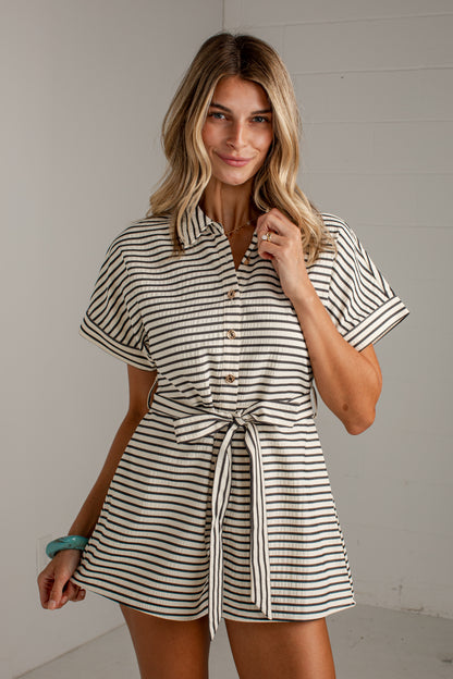 A woman modeling a short-sleeve collared romper with thin black horizontal stripes on a cream background, featuring a button-front bodice and a self-tie waist belt.