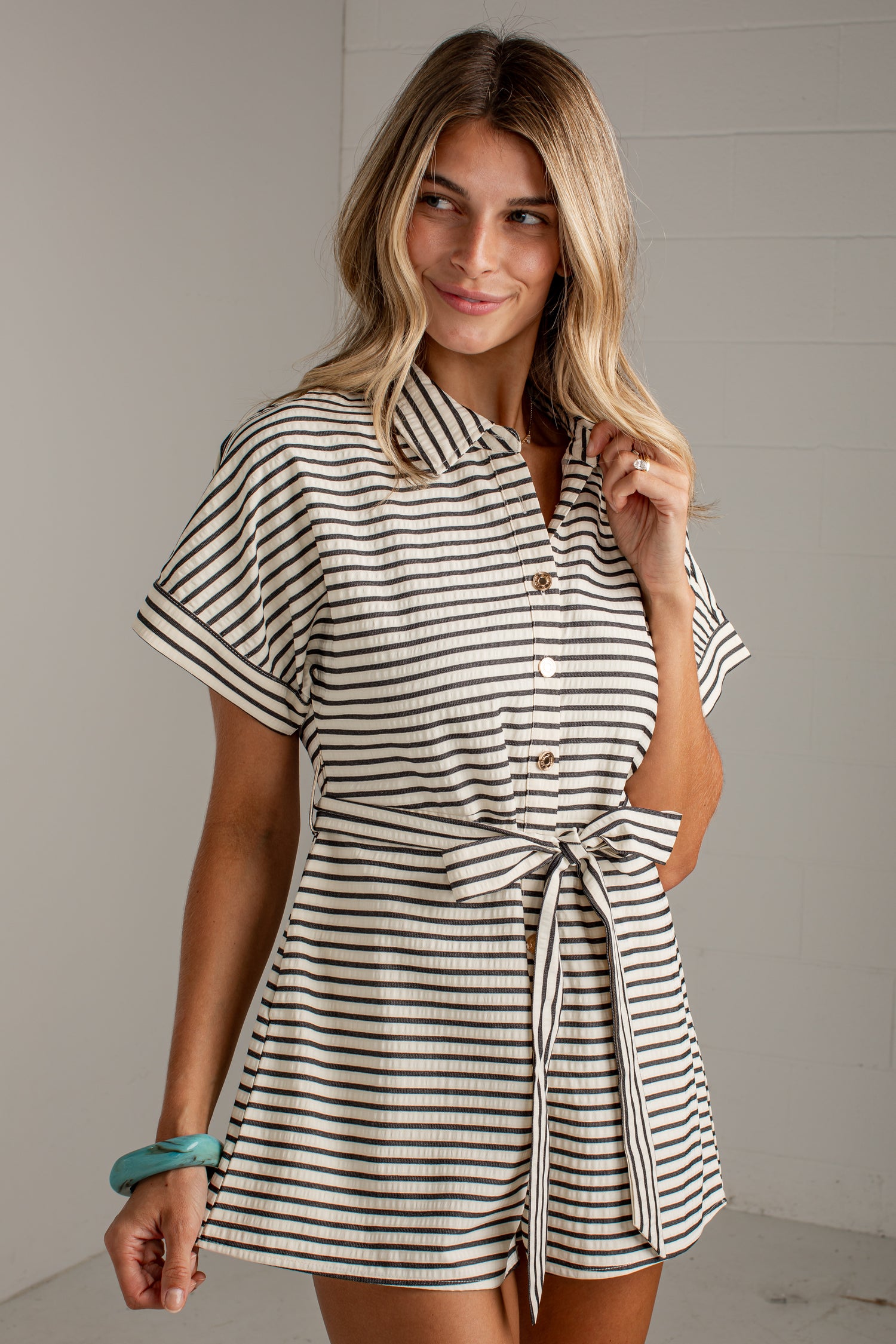 A woman modeling a short-sleeve collared romper with thin black horizontal stripes on a warm off-white base, featuring a button-front neckline and a self-tie waist belt.