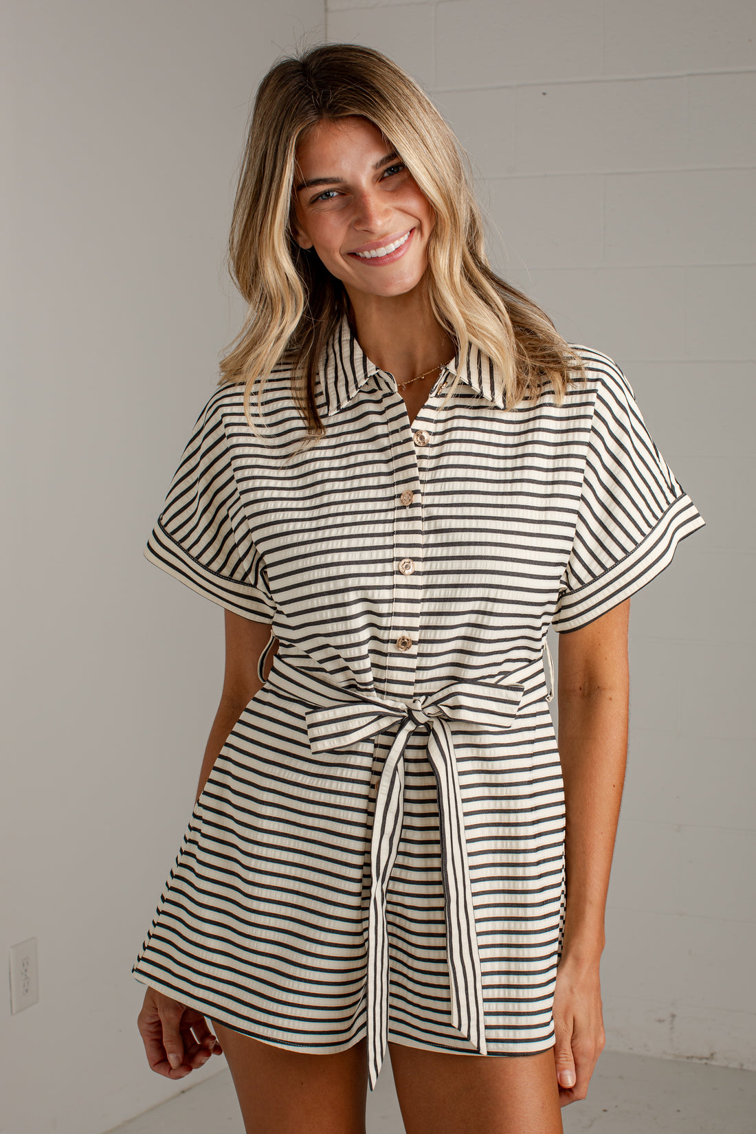 A woman modeling a short-sleeve collared romper with thin black and off-white horizontal stripes, featuring a button front and a self-tie belt at the waist.