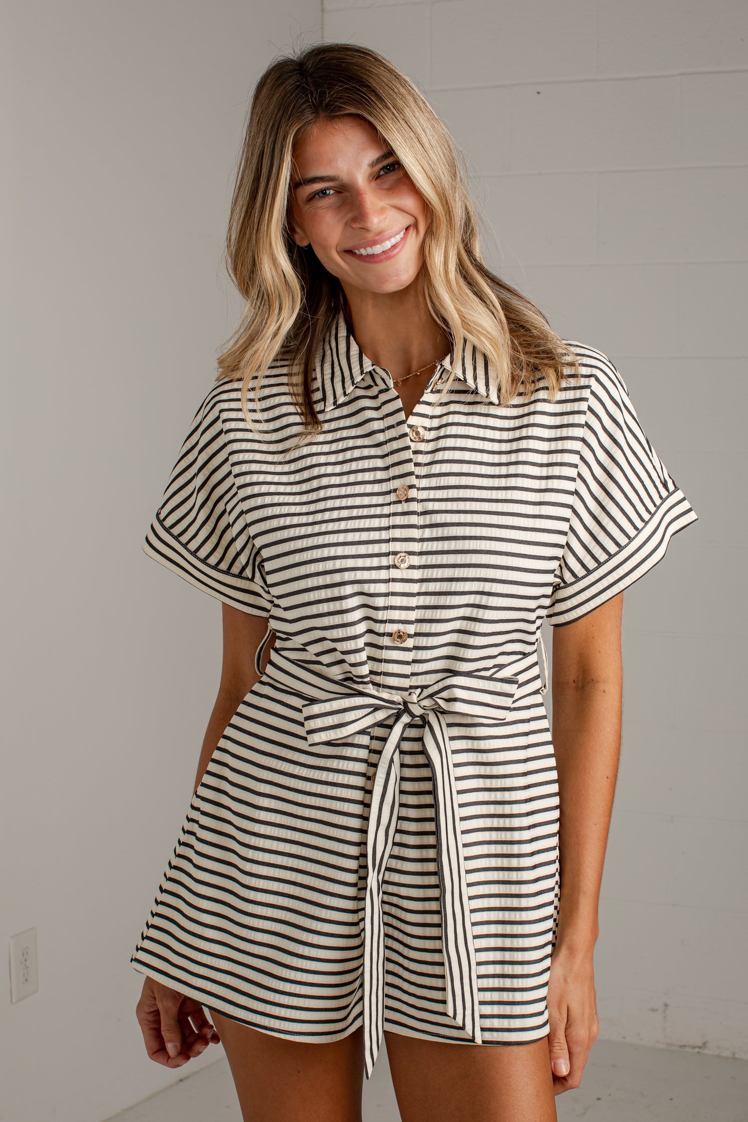 A woman modeling a short-sleeve collared romper with thin black and off-white horizontal stripes, featuring a button front and a self-tie belt at the waist.
