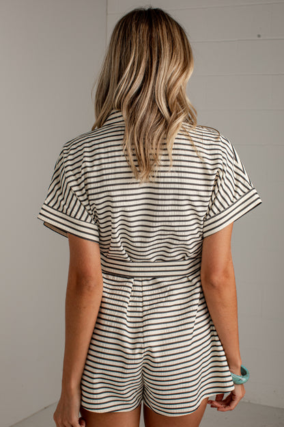 A woman viewed from behind wearing a short-sleeve collared romper with thin horizontal black stripes on an off-white base, standing against a light neutral wall.