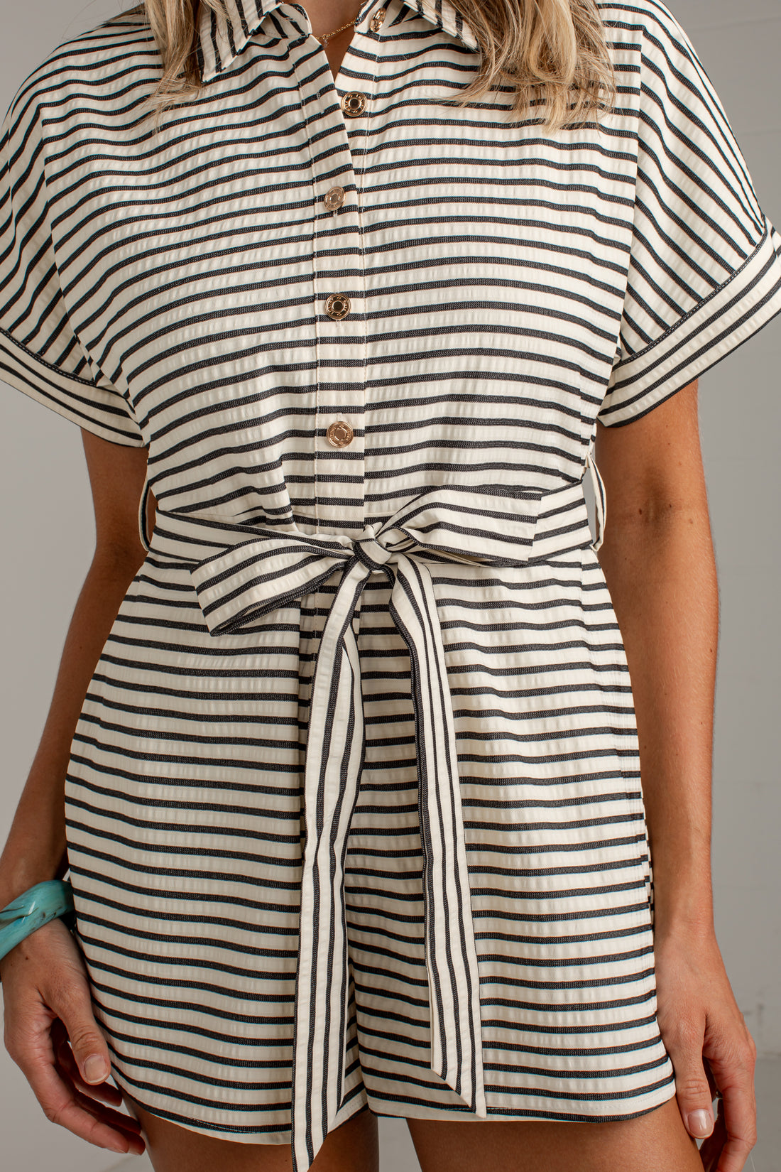 A close-up of a model wearing a short-sleeve collared romper with black and off-white horizontal stripes, featuring a button-down front and a matching tie belt at the waist.