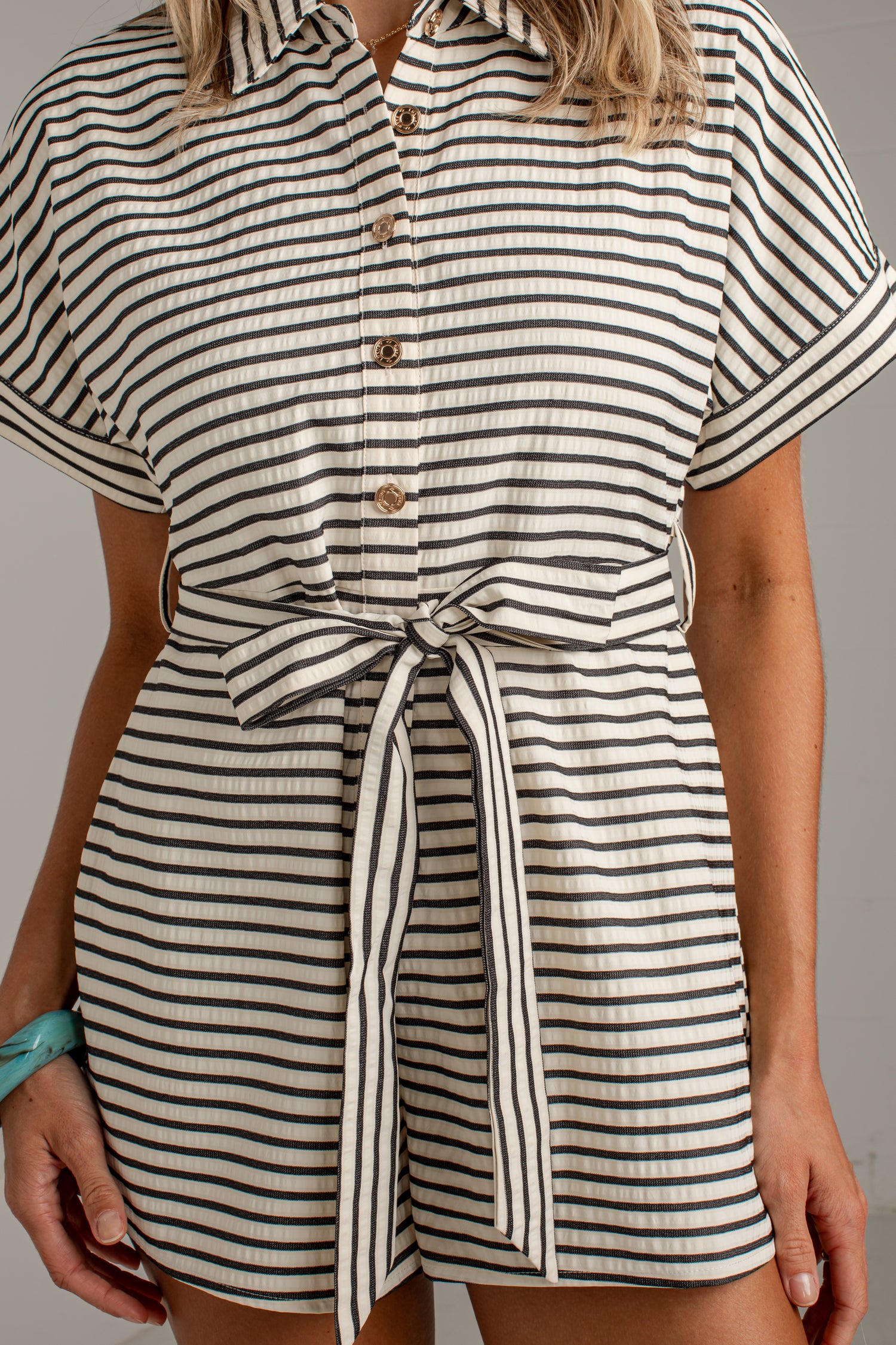 A close-up of a model wearing a short-sleeve collared romper with black and off-white horizontal stripes, featuring a button-down front and a matching tie belt at the waist.