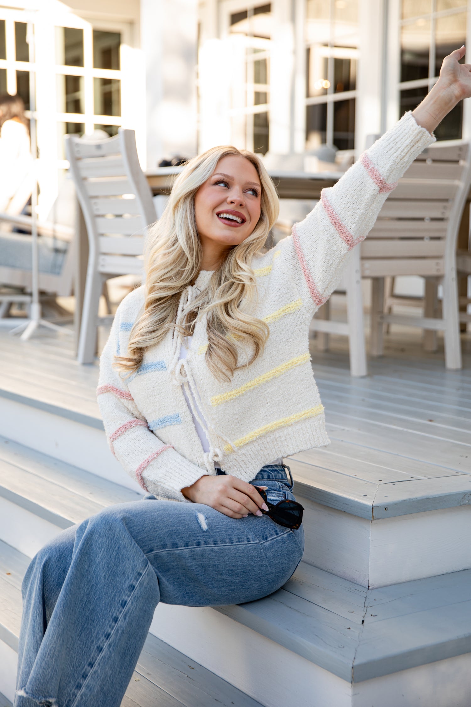 Woman sitting on steps wearing a colorful sweater and jeans, with a blurred outdoor setting.