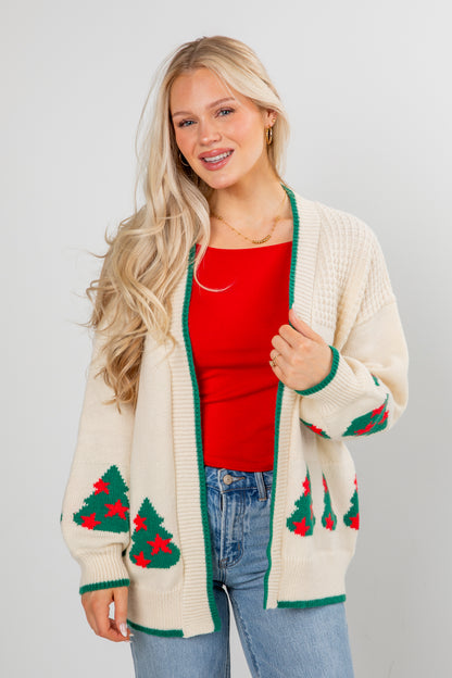Woman wearing a cream cardigan with Christmas tree patterns, red top, and blue jeans on a light gray background