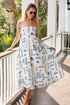 Woman in a white dress with blue patterns standing on a wooden deck.