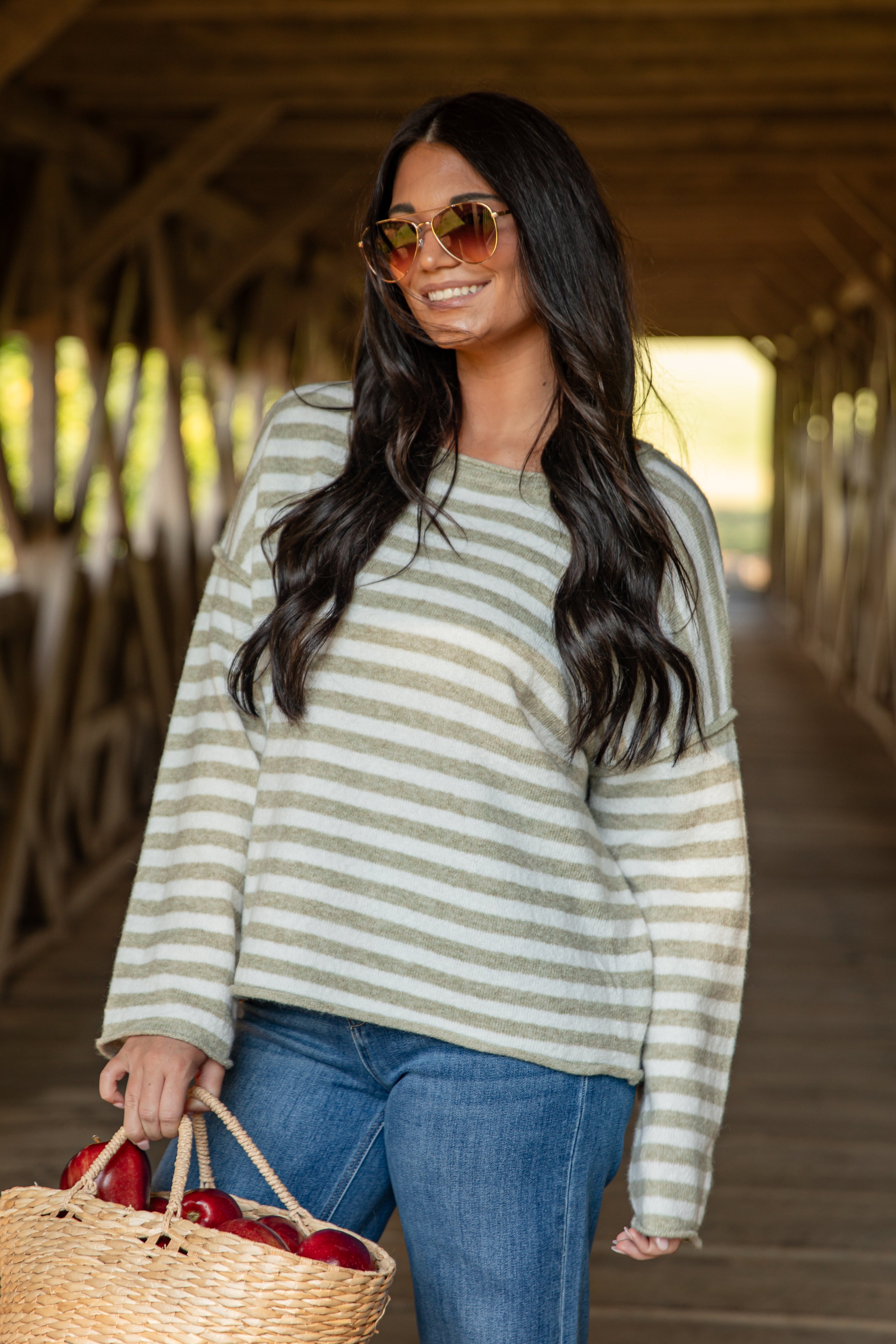 Woman wearing a striped sweater and sunglasses, holding a basket with cherries, standing on a wooden bridge.