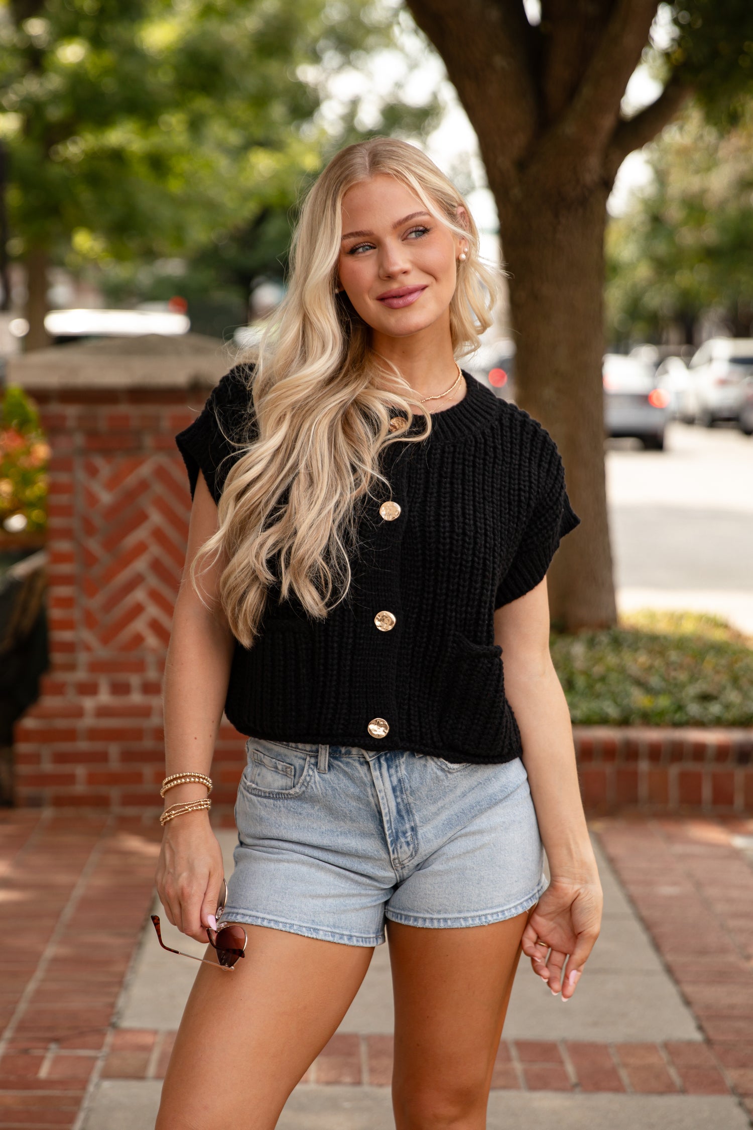 Woman wearing a black top with gold buttons and denim shorts outdoors.