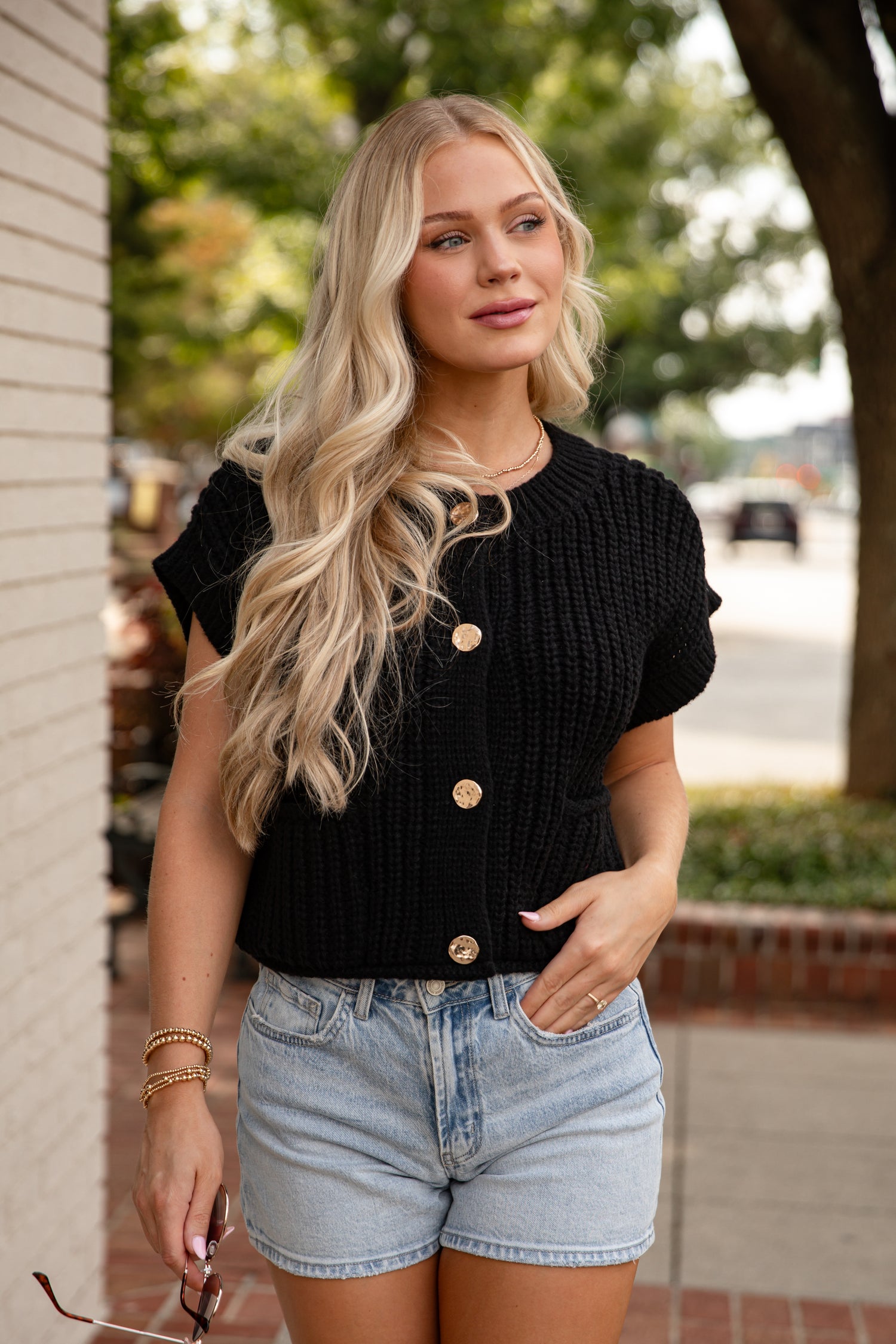 Woman wearing a black cardigan with gold buttons and denim shorts outdoors.