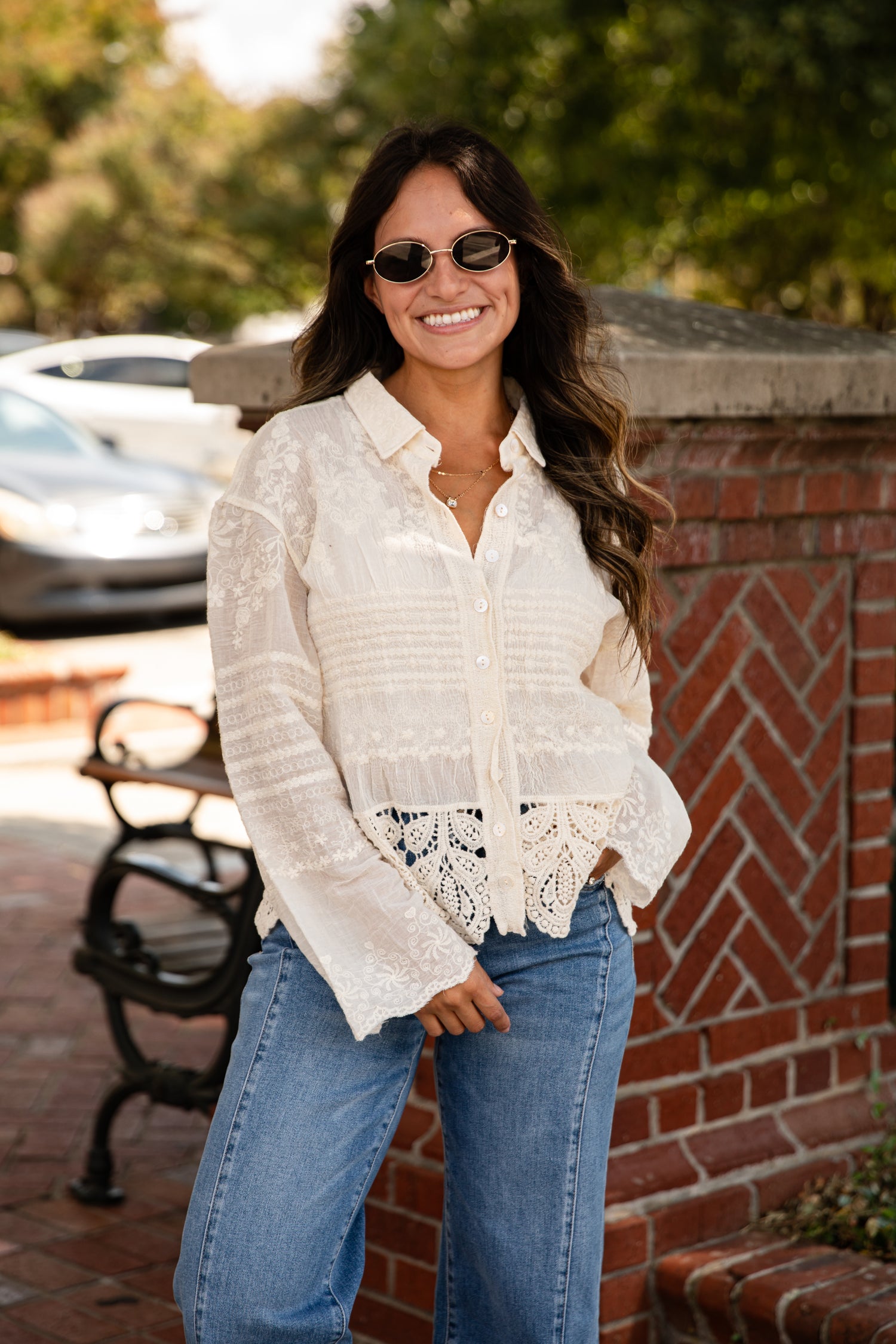 Woman wearing a white blouse with lace details and blue jeans standing outdoors near a brick wall.