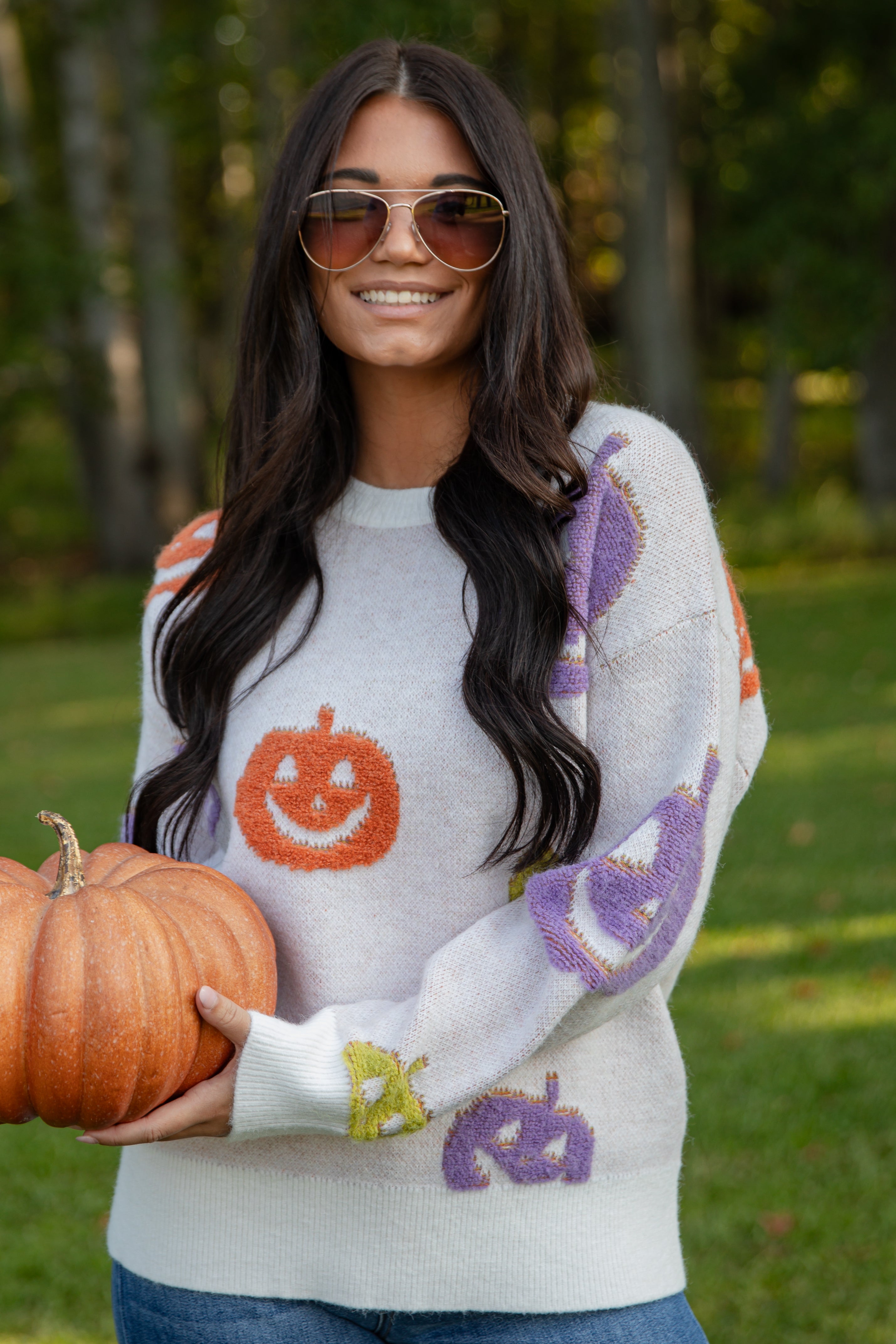 Woman wearing a Halloween-themed sweater holding pumpkins outdoors.