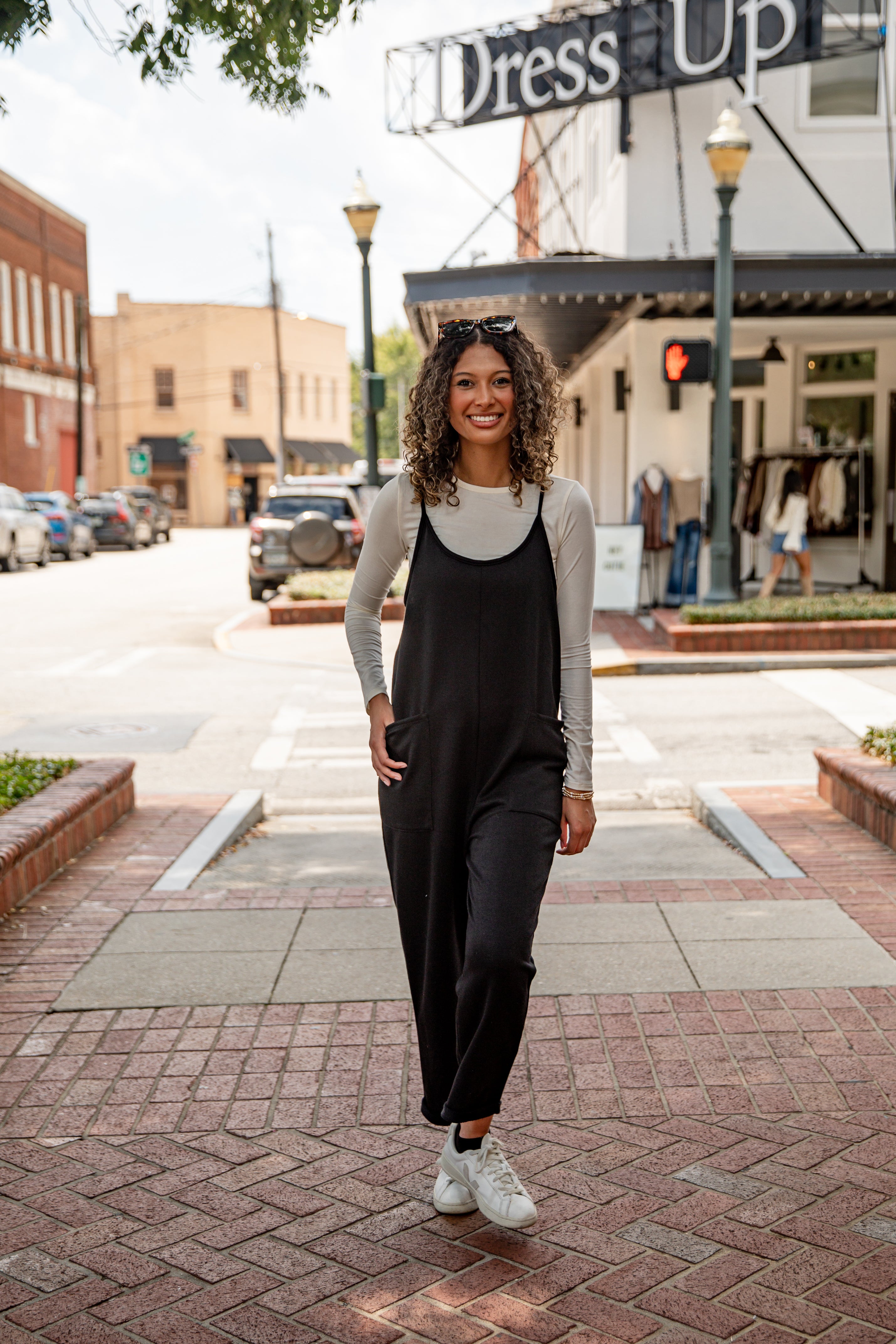 Woman in black jumpsuit and white sneakers standing on a city street with &