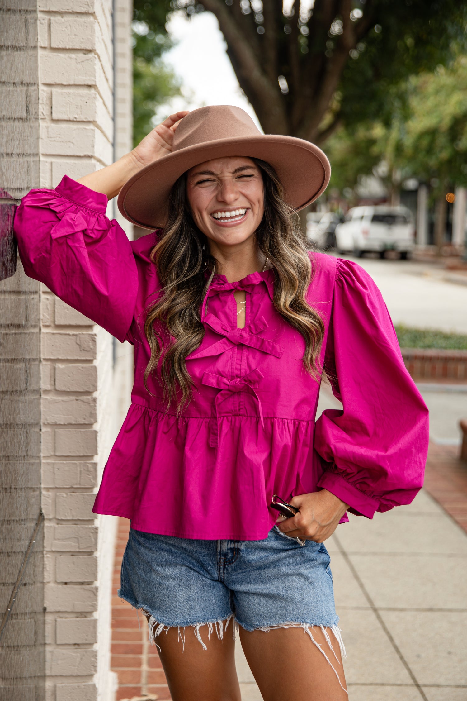 Woman wearing a bright pink blouse, denim shorts, and a beige hat, standing against a brick wall.