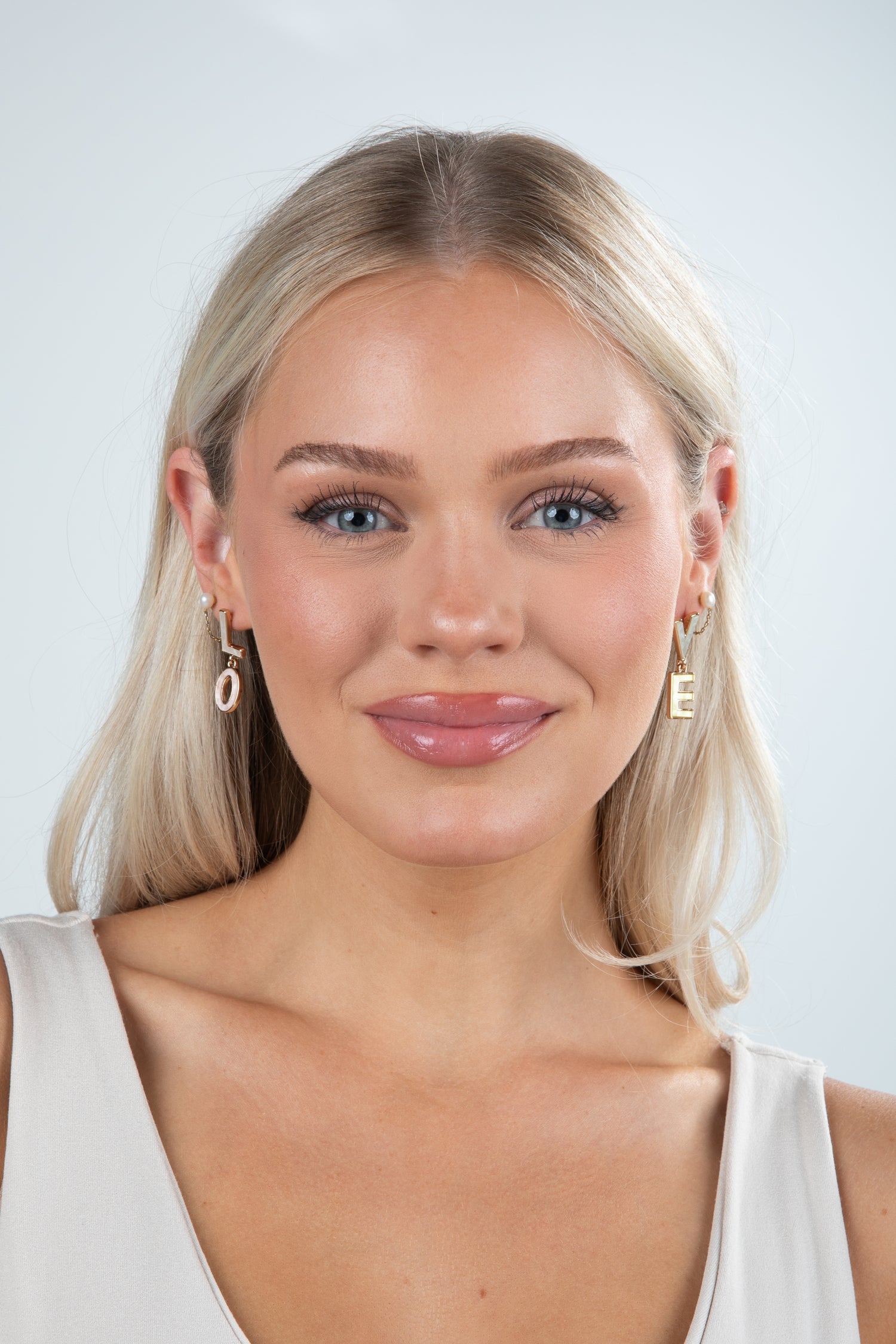 Woman with blonde hair and earrings wearing a white top against a light gray background