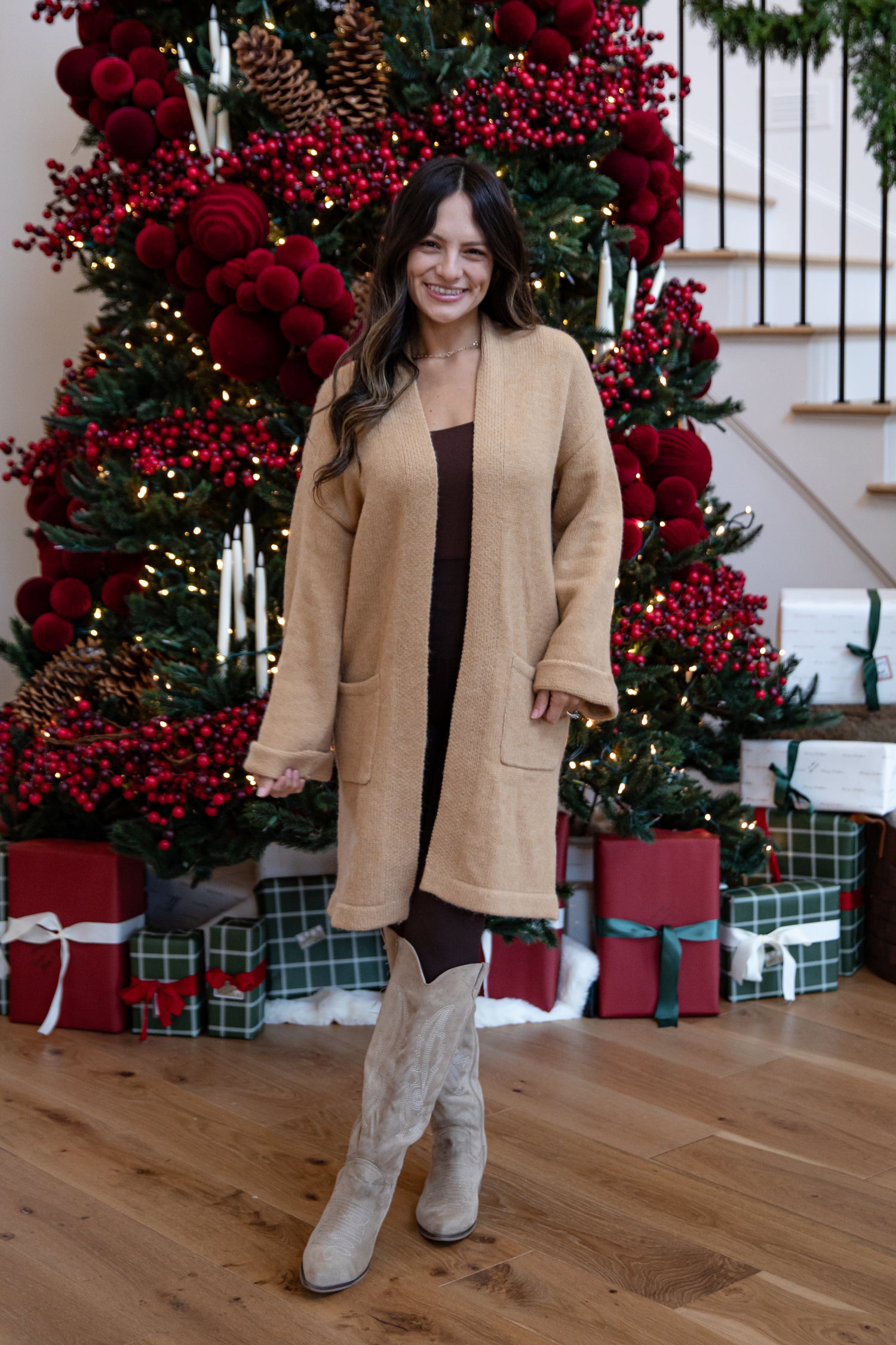 Woman in a beige coat standing in front of a decorated Christmas tree with red berries and lights.