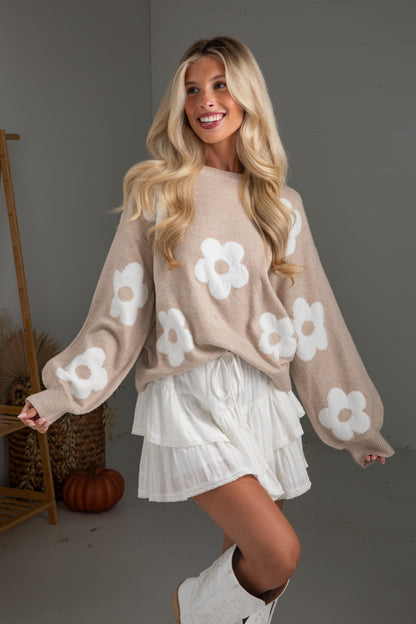 Woman wearing a beige sweater with white floral patterns and a white skirt.