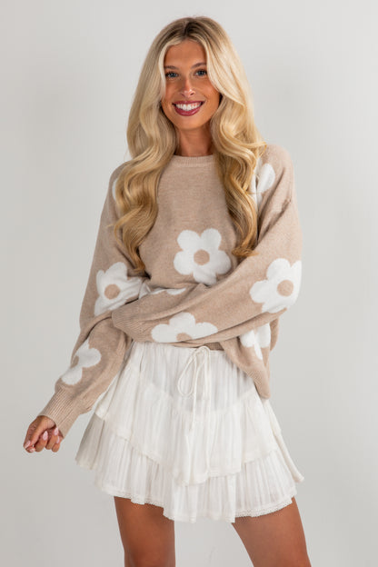 Woman wearing a beige sweater with white floral patterns and a white skirt on a plain background