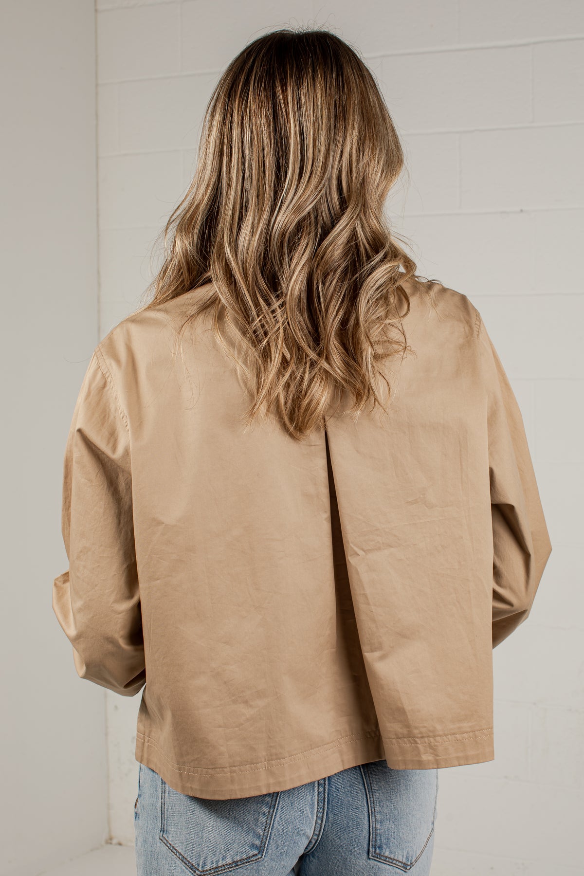 A woman viewed from behind wearing a light tan utility-style jacket and light blue jeans, standing against a white brick wall.
