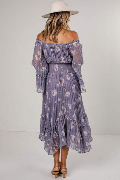 A woman shown from the back wearing a lavender off-the-shoulder midi dress with a white floral print, tiered ruffle sleeves, and a matching ruffled skirt. She is also wearing a wide-brimmed cream hat and brown strappy heels against a plain light gray studio background.
