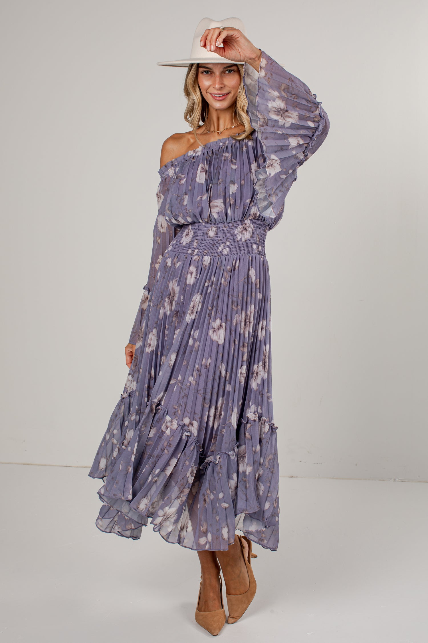 A woman modeling a long-sleeve off-the-shoulder midi dress with a purple and white floral print, paired with a white wide-brim hat and tan pointed-toe heels, set against a plain light gray studio background.