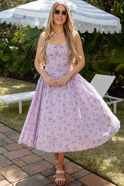 Woman in a purple floral dress standing under a striped umbrella in a garden setting.