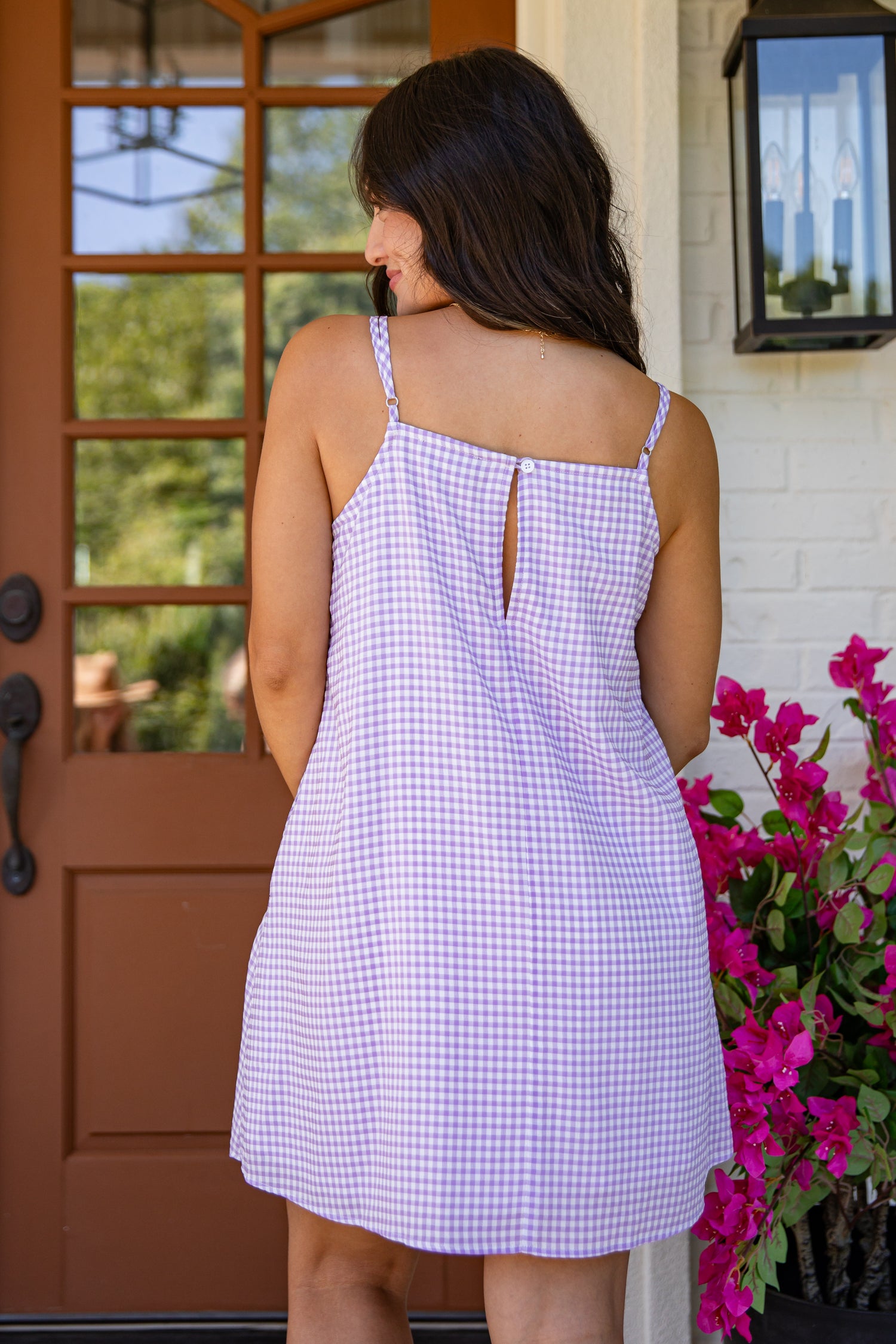 Woman wearing a checkered dress standing in front of a door with flowers to her right.