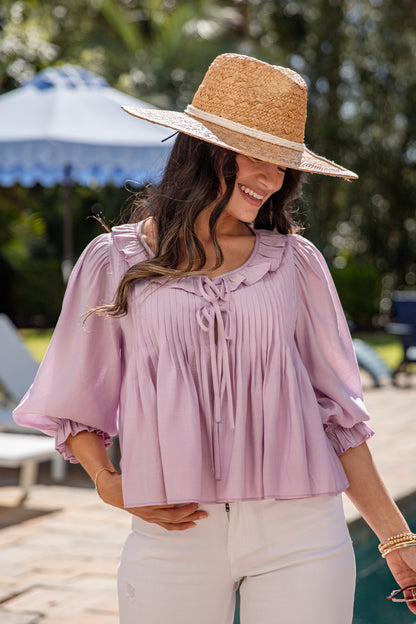 Woman wearing a straw hat and light purple blouse with white pants outdoors.