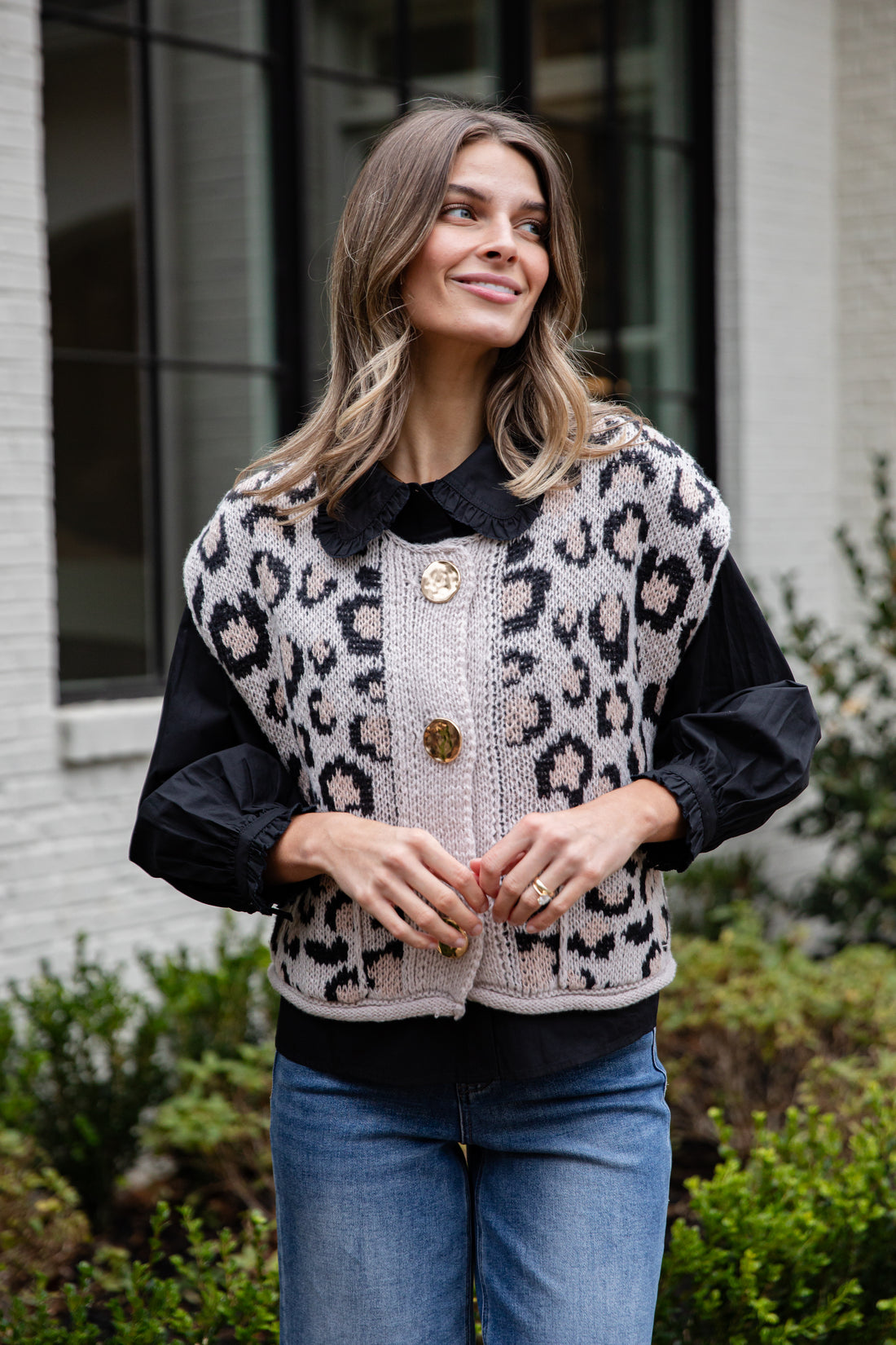 Woman wearing a leopard print vest over a black shirt with blue jeans, standing outdoors.