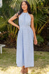 Woman in a light blue dress standing in a garden with palm trees and a lounge chair.