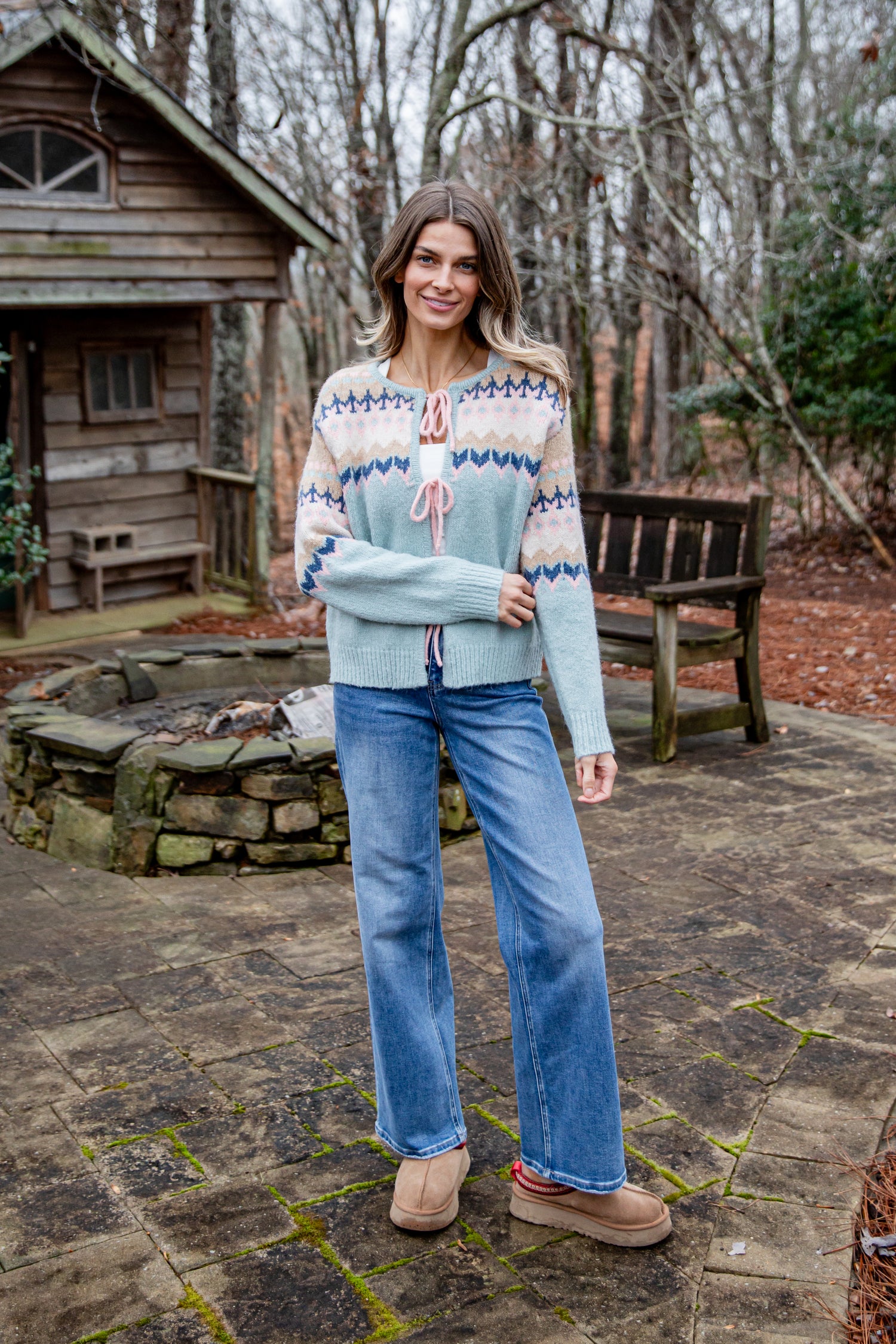 Woman wearing a patterned sweater and jeans standing outdoors near a wooden cabin.