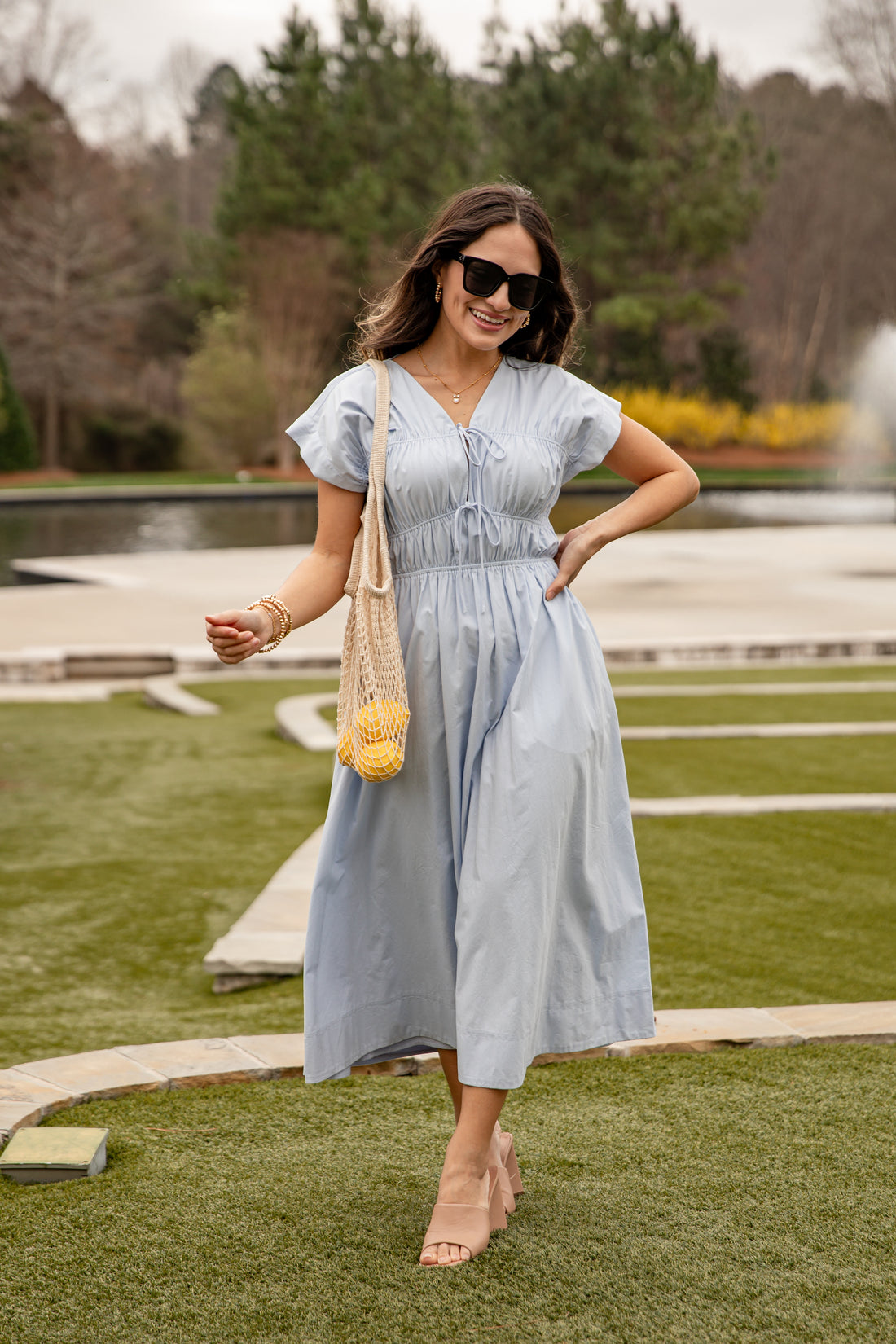 Woman in a light blue dress standing outdoors with a fountain and greenery in the background