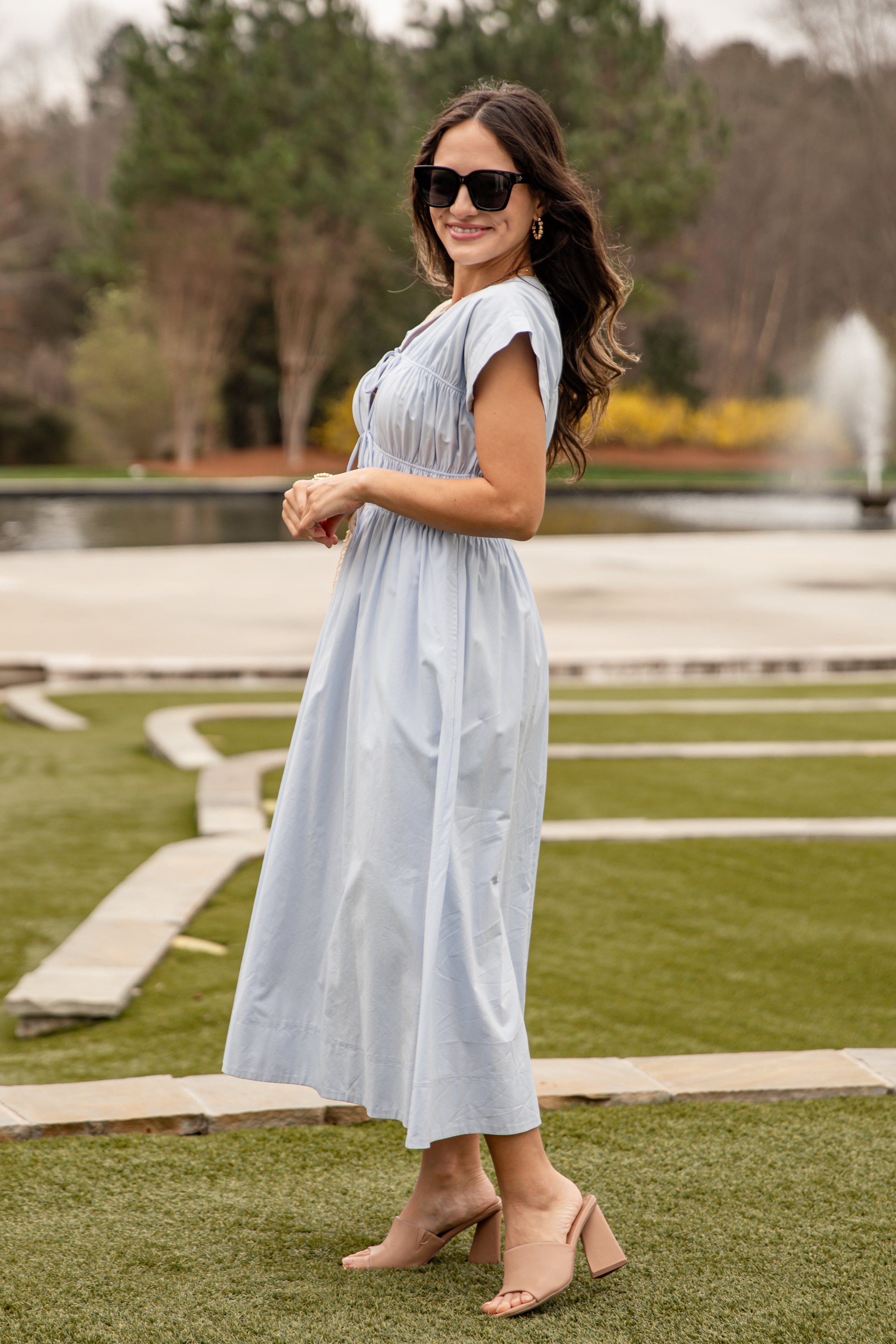 Woman in a light blue dress standing in a park with trees and a fountain in the background.