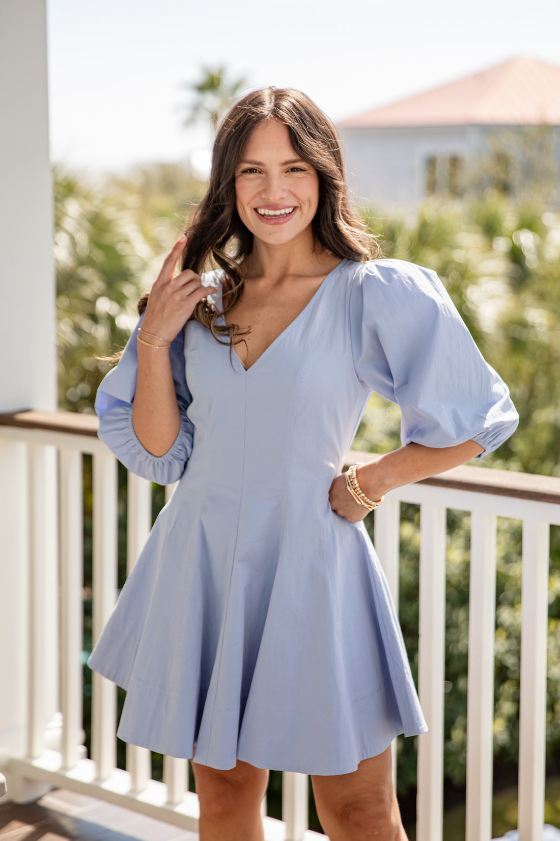 Woman in a light blue dress standing on a balcony with greenery in the background