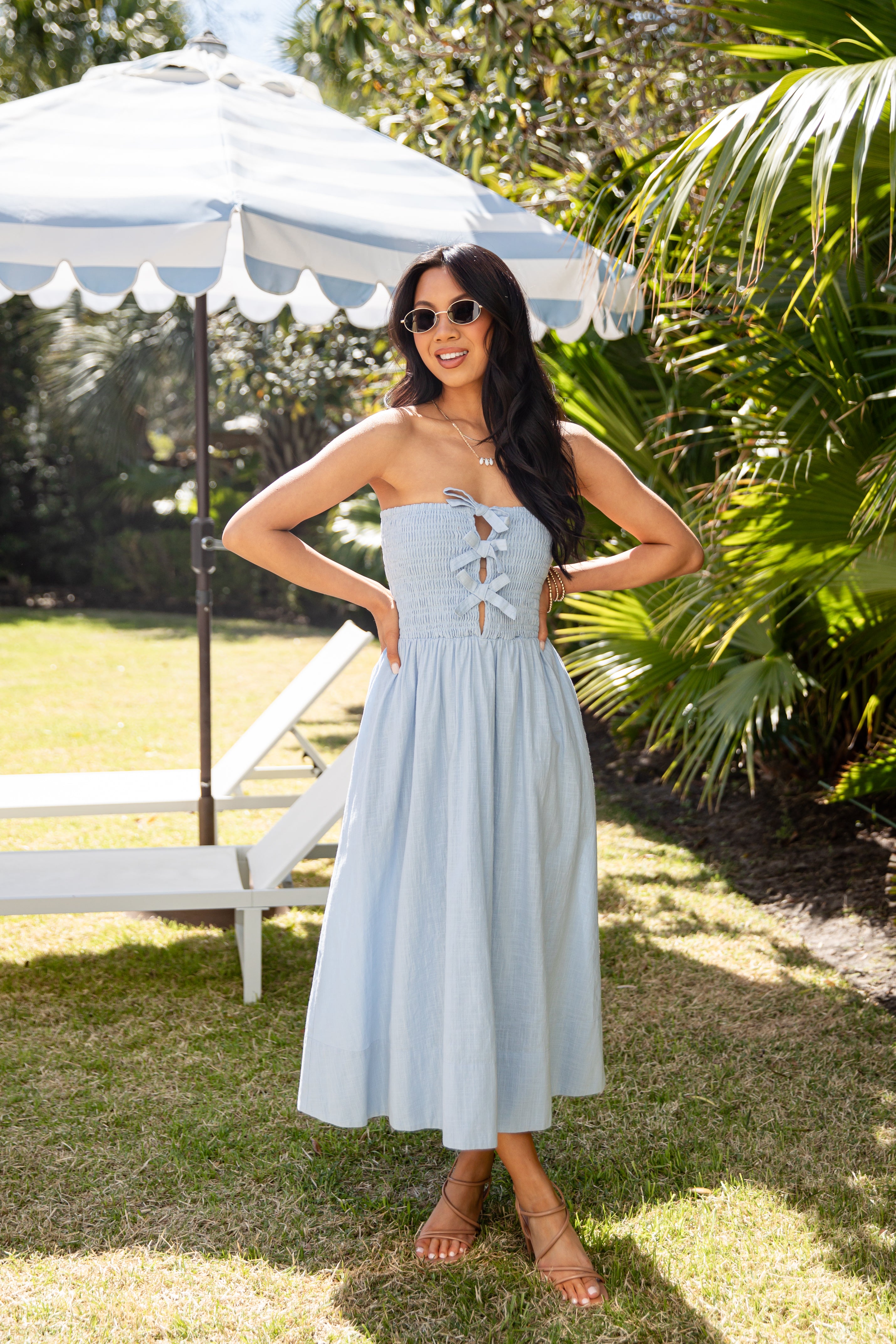 Woman in a light blue dress standing outdoors with greenery and a white umbrella in the background