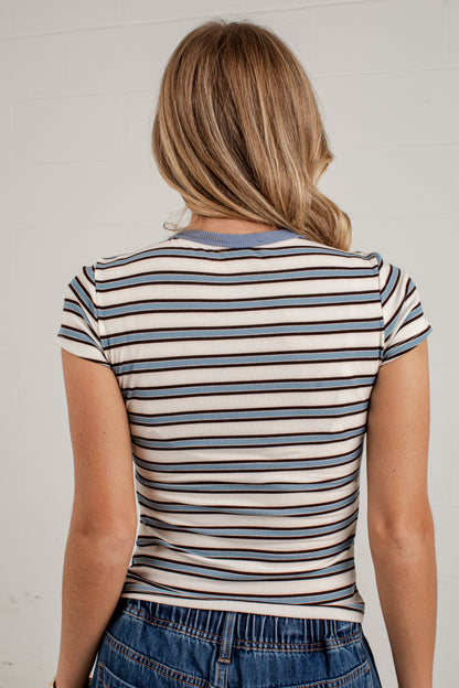 A woman viewed from behind wearing a fitted short-sleeve striped top with horizontal navy, blue, and beige stripes, paired with blue jeans against a light neutral wall.