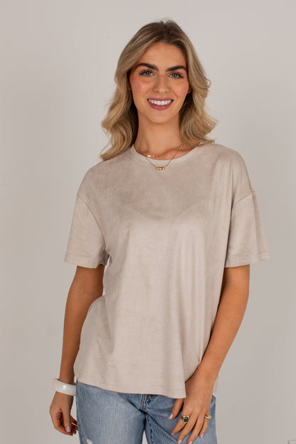 A woman with blonde hair wearing a light beige short-sleeve crewneck t-shirt and blue jeans, standing against a neutral light gray background.