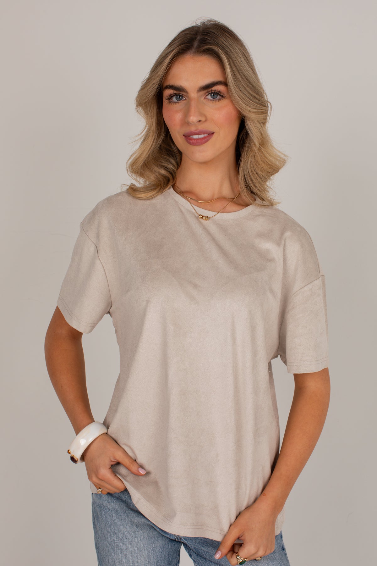 A woman wearing a light beige short-sleeve crewneck top and blue jeans, standing against a neutral light gray background.